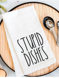 White towel with 'STUPID DISHES' text on a wooden surface with measuring spoons.