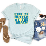 Light blue t-shirt with 'Life is Better at the Beach' text, flip-flops, sunglasses, shorts, and beach accessories on a white background.